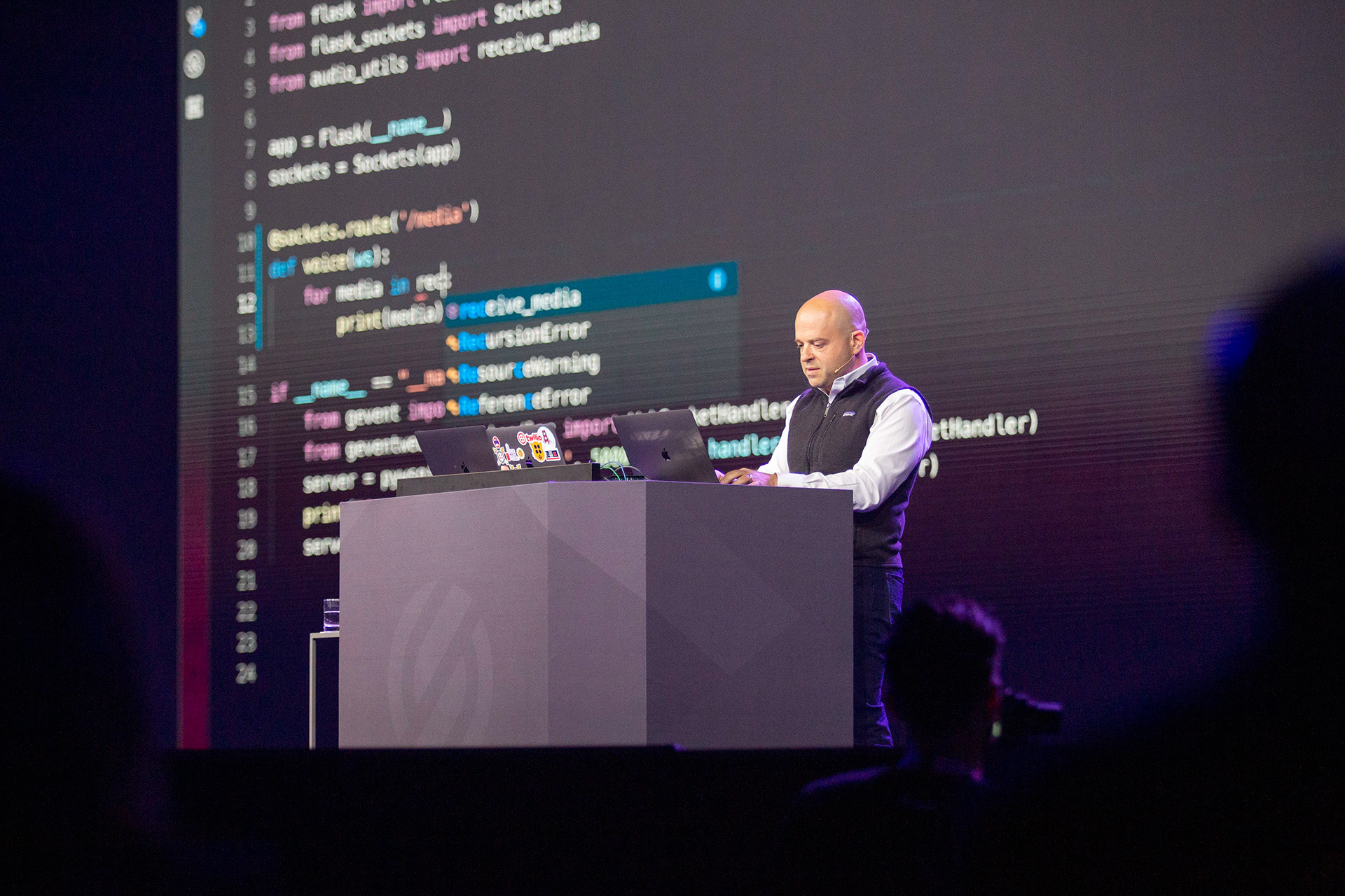 Speaker live-coding on stage with Python code displayed on large screen behind him
